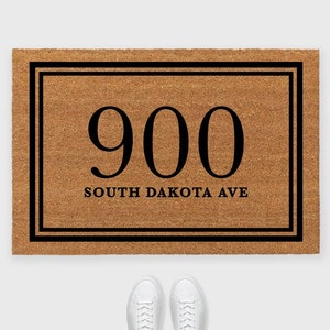 May include: A brown coir doormat with a black border and double black frame. The number 900 is printed in black inside the frame, along with the text "SOUTH DAKOTA AVE".