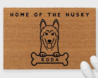 May include: A brown doormat with the text "Home of the Husky" and a black and white illustration of a husky dog holding a bone with the name "Koda" written on it.
