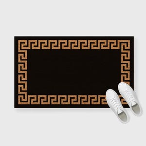 May include: Black rectangular doormat with a Greek key border in a light brown color. Two white sneakers are placed near the mat. The doormat is a home decor item.