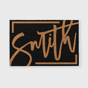 May include: A black doormat with a gold frame and the word "Smith" written in a cursive font.
