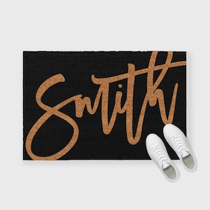 K&ouml;nnte beinhalten: Schwarze Fu&szlig;matte mit dem Wort "Smith" in goldener Kursivschrift.
