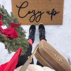 May include: A woman wearing black pants, a white fuzzy jacket, and red and black socks. She is wearing black and white boots with fur trim. She is holding a brown quilted handbag with a gold chain strap. The woman is standing in front of a brown doormat with the words "Come in & Cozy up" written in black.