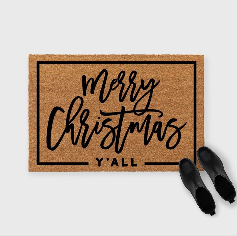 May include: A rectangular doormat with the words "Merry Christmas Y'all" in black script. The mat is brown with a black border. Two black ankle boots are placed to the right of the mat.