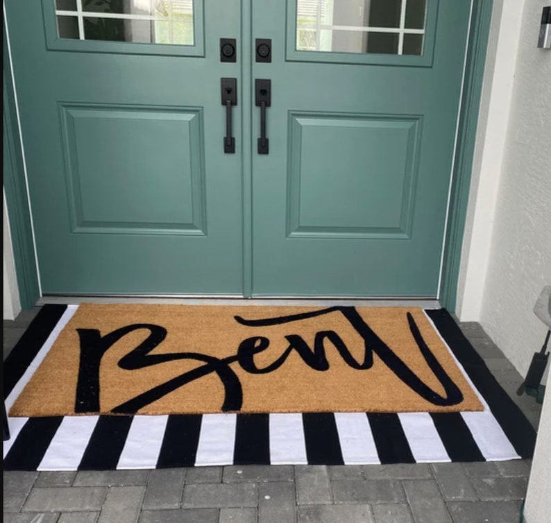 May include: A brown coir doormat with the word "Bent" written in black script. The doormat is placed on a black and white striped rug.