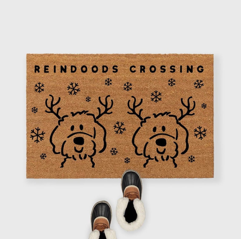 May include: A brown coir doormat with the text "REINDOODS CROSSING" in black lettering. Two black and white illustrations of dogs with antlers are on the mat, surrounded by snowflakes.