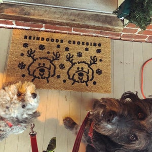 May include: A brown doormat with the text "Reindeer Crossing" and two black reindeer silhouettes. Two dogs, one gray and one black, are standing on the doormat.