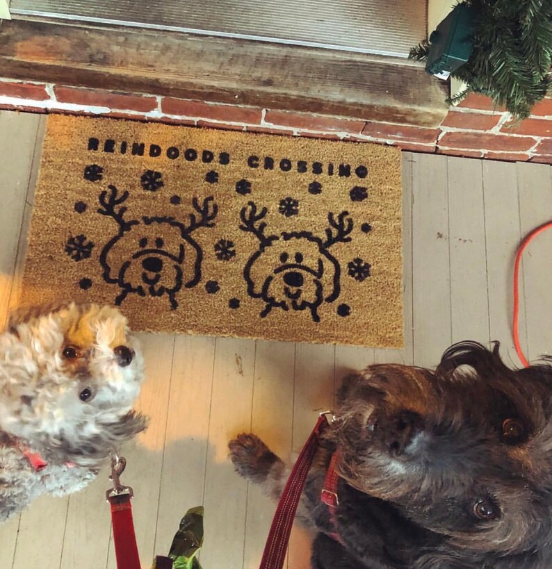 May include: A brown doormat with the text "Reindeer Crossing" and two black reindeer silhouettes. Two dogs, one gray and one black, are standing on the doormat.