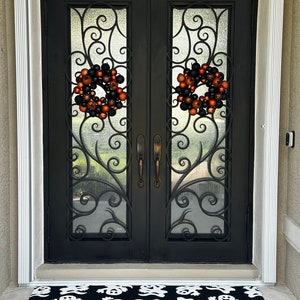 Puede incluir: Dos puertas dobles negras con un dise&ntilde;o de hierro forjado ornamentado y paneles de vidrio. Dos coronas de Halloween con adornos naranja y negro cuelgan de las puertas. Una alfombra de bienvenida negra y blanca con un patr&oacute;n de fantasma est&aacute; frente a las puertas.