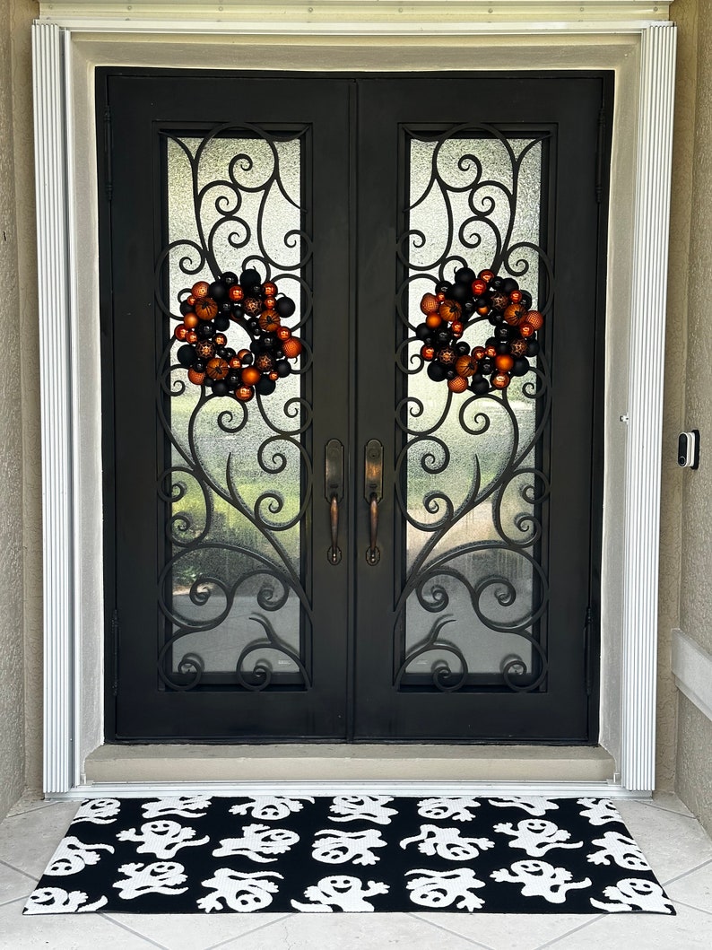 Puede incluir: Dos puertas dobles negras con un dise&ntilde;o de hierro forjado ornamentado y paneles de vidrio. Dos coronas de Halloween con adornos naranja y negro cuelgan de las puertas. Una alfombra de bienvenida negra y blanca con un patr&oacute;n de fantasma est&aacute; frente a las puertas.