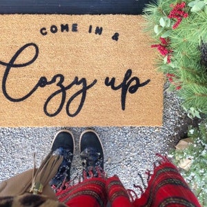 May include: A brown coir doormat with the text "Come in & Cozy up" in black script. A person is standing on the mat wearing black boots and a red and black plaid scarf.