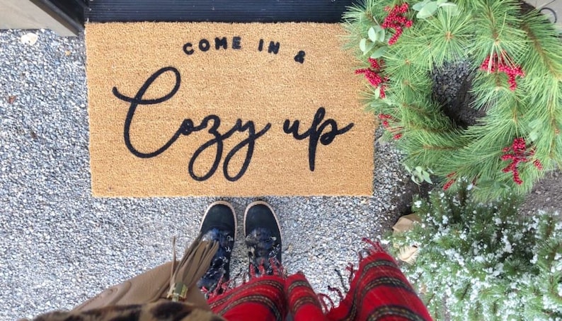 May include: A brown coir doormat with the text "Come in & Cozy up" in black script. A person is standing on the mat wearing black boots and a red and black plaid scarf.
