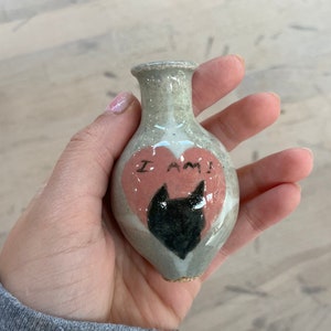 May include: A small, light gray ceramic vase with a pink heart painted on the front. The heart has a black cat silhouette inside and the words "I AM" written on it.