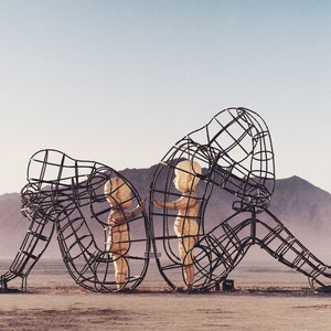 Könnte beinhalten: Eine große Metallskulptur aus zwei Figuren, eine sitzt und die andere steht, beide scheinen kleinere Figuren in ihren Rahmen zu halten. Die Skulptur ist vor dem Hintergrund von Bergen und einer staubigen Wüstenlandschaft platziert.