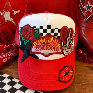 May include: A red and white trucker hat featuring a central embroidered patch that reads HOT GIRLS READ SPICY BOOKS in red and yellow flames. The hat includes additional patches of a red rose, a black and white checkered pattern, a red heart, and a red lip print.