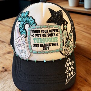 Könnte beinhalten: Eine schwarz-weiße Trucker-Mütze mit Mesh-Rückseite. Die Mütze hat türkisfarbene Verzierungen und Aufnäher, darunter ein Stern, ein Blitz und ein Aufnäher mit dem Text "DRINK YOUR COFFEE PUT ON SOME TURQUOISE AND HANDLE YOUR SH*T."