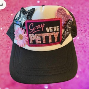 May include: A black and white trucker hat with a curved brim. The hat features a patch that reads "Sorry We're Petty" in pink and black. It is decorated with a pink flower, a star, a heart, and a lightning bolt.
