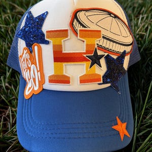 May include: A blue and white trucker hat with a large orange and yellow letter "H" and a baseball stadium patch. The hat features blue sequin stars, an orange star, and the text "Let's Go!".