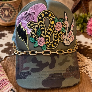 May include: A camouflage print trucker hat with a gold chain and decorative patches. The patches include a snake, roses, a lightning bolt, and a hand gesture. The hat has a curved brim and a mesh back.