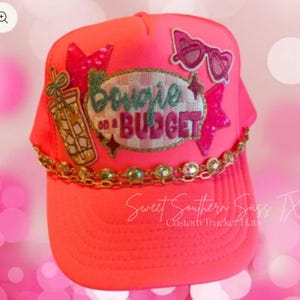 Könnte beinhalten: Hellrosa Trucker-Cap mit Mesh-Rückseite und gebogenem Schirm. Die Kappe hat eine goldene Kette, Aufnäher mit Sonnenbrillen, einem Getränk und dem Schriftzug "Bougie on a Budget". Der Text "Sweet Southern Sass TX" ist ebenfalls sichtbar.