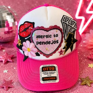 May include: A white and hot pink trucker hat featuring a central sequined heart patch with the text 'allergic to pendejos'. The hat is decorated with embroidered patches including a red lip, a pink flower, a diamond, and a black lightning bolt.