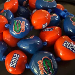 May include: A collection of orange and blue beads, some with the text "2025" and others with a Gator logo. The beads are strung together with blue ribbon, creating a festive look. The Gator logo features a green alligator.