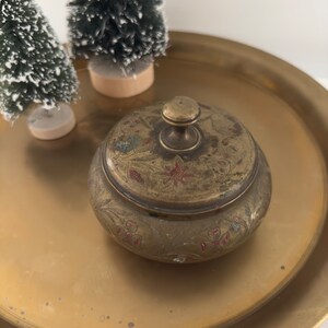 May include: A vintage brass trinket box with a lid, adorned with floral and leaf engravings and red accents. The box sits on a gold-toned tray, with two small Christmas trees in the background. The box has a round shape and a small knob on the lid.