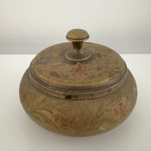 May include: A vintage brass trinket box with a lid. The box is round with floral and leaf engravings and red accents. The lid has a small knob for lifting. The brass has a tarnished, aged appearance.