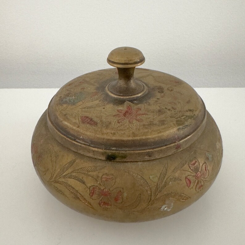 May include: A vintage brass trinket box with a lid. The box is round with floral and leaf engravings and red accents. The lid has a small knob for lifting. The brass has a tarnished, aged appearance.