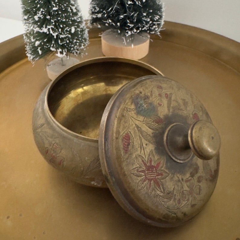 May include: An antique brass lidded bowl with floral and leaf engravings and red accents. The lid features a round knob and is partially open, revealing the bowl's interior. The bowl sits on a gold-toned tray.