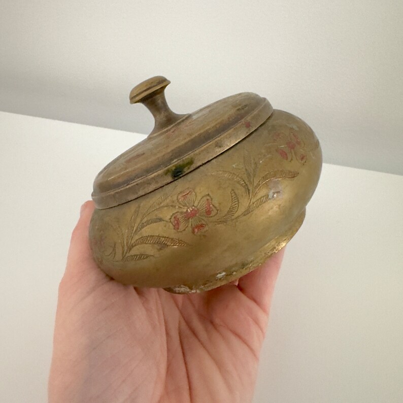 May include: A vintage brass trinket box with a lid. The box is oval-shaped and features etched floral designs with red accents. The lid has a small knob for easy opening. The brass has a warm, aged patina.