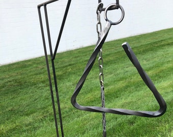 Puede incluir: Un carillón triangular de metal negro que cuelga de una cadena unida a un soporte de metal negro. El soporte tiene una base triangular y un solo poste vertical.