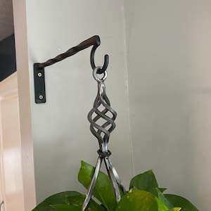 Op de afbeelding: Een zwarte metalen plantenhanger met een gedraaide arm en een decoratieve kooi aan het einde. De hanger is aan een muur bevestigd en een groene plant hangt aan de kooi.
