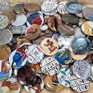 May include: A collection of round, colorful buttons featuring images of Egyptian hieroglyphics, gods, and symbols. The buttons are arranged in a random pattern on a brown surface.