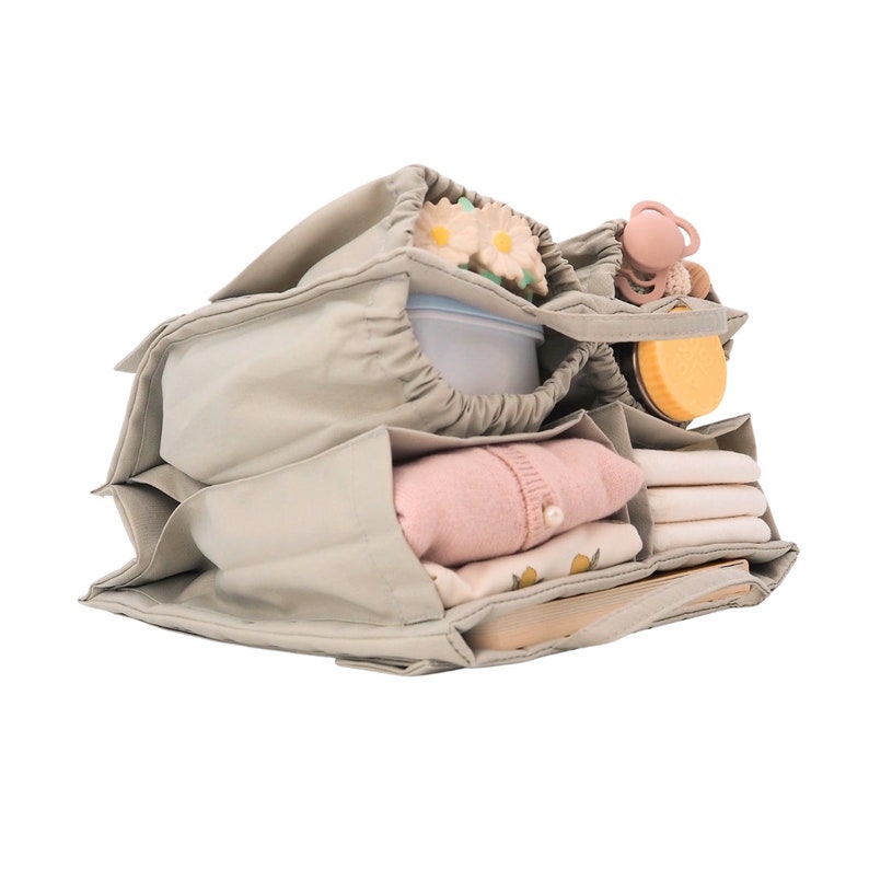 May include: A gray canvas organizer with multiple compartments, filled with baby items like a pink sweater, a white onesie, a yellow bottle, and a pacifier.