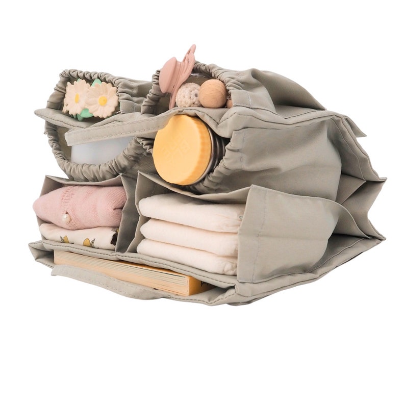 May include: A gray canvas organizer with multiple compartments filled with baby items, including a bottle, diapers, a pacifier, and a book.