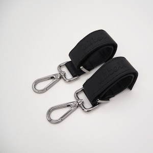 Op de afbeelding: Twee zwarte leren banden met zilveren metalen clips. De banden hebben het woord "little" erop gegraveerd.