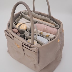 May include: A light gray tote bag with a Prada logo and a zippered pocket. The bag is open and filled with various items, including a pink sweater, a white cloth, and a small white box.