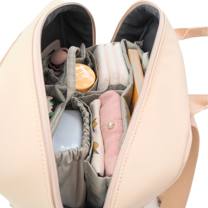 May include: A light pink diaper bag with multiple compartments and pockets. The bag is open and filled with baby supplies, including diapers, wipes, a bottle, and a small toy.