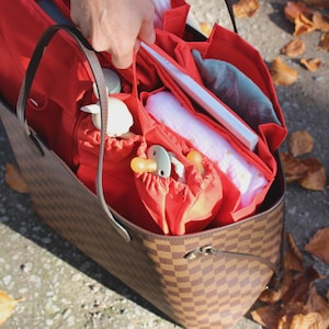 Peut inclure: Un sac fourre-tout à carreaux marron et beige contenant un insert organisateur en tissu rouge vif. L'insert comporte plusieurs compartiments contenant divers objets, dont un livre blanc, une sucette claire et d'autres petits accessoires.