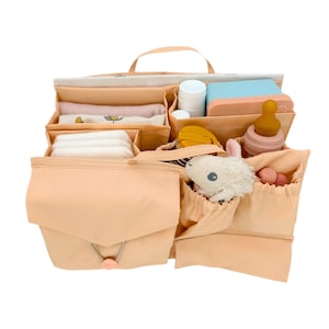 May include: A peach-colored diaper bag organizer with multiple compartments and pockets. The organizer is filled with baby essentials, including diapers, wipes, a bottle, a stuffed animal, and a small toy.