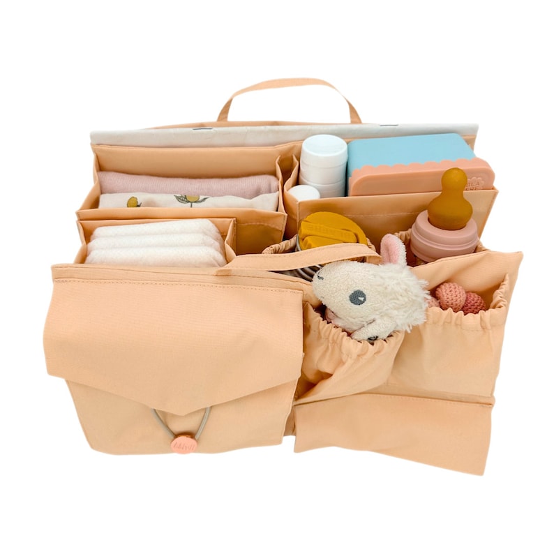 May include: A peach-colored diaper bag organizer with multiple compartments and pockets. The organizer is filled with baby essentials, including diapers, wipes, a bottle, a stuffed animal, and a small toy.