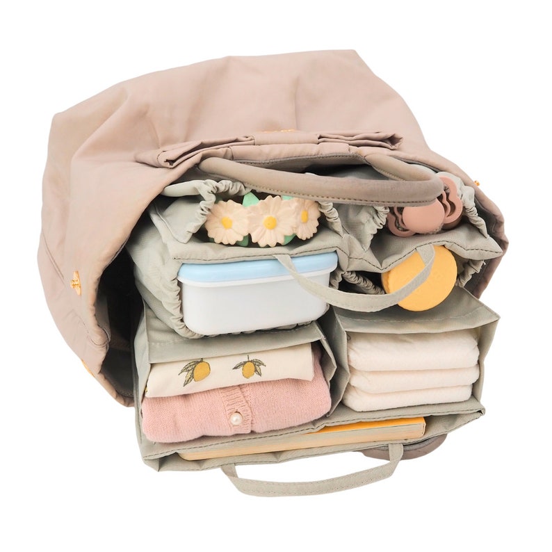 May include: A beige tote bag with multiple compartments and pockets. The bag is filled with various items, including a lunchbox, a small container, a baby bottle, and a few toys. The bag is open and the contents are visible.