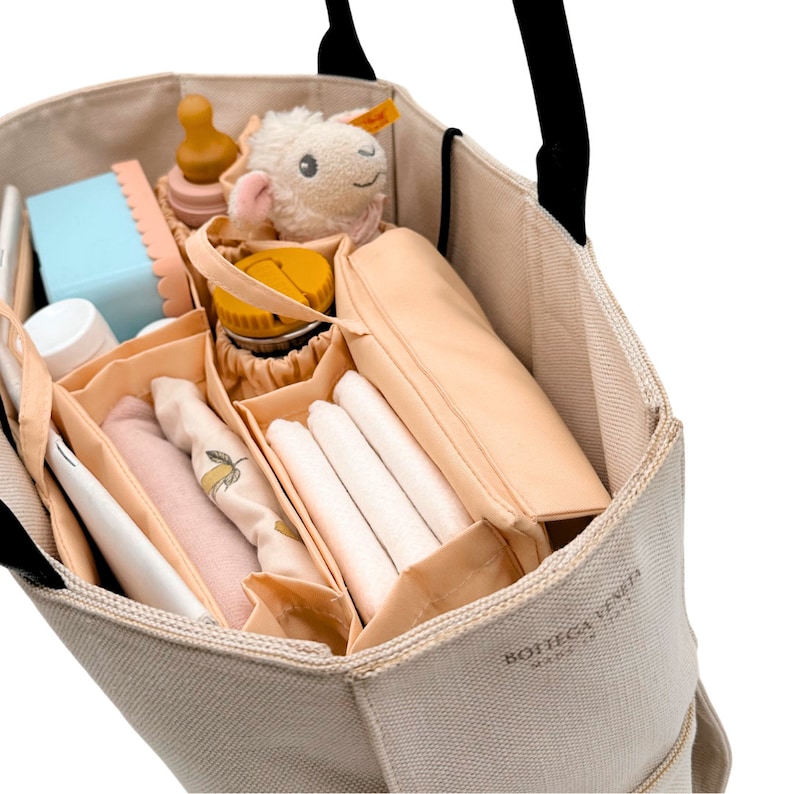May include: A beige canvas tote bag with a black handle and a beige and brown organizer insert. The organizer has multiple compartments and is filled with various items, including a stuffed animal, a water bottle, and a diaper.
