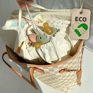 Può includere: Una borsa tote a scacchi beige e bianca contenente sacchetti di stoffa bianca e diversi ciucci in silicone nei toni del verde salvia, rosa polvere e giallo ambra. È attaccata un'etichetta beige con il testo ECO e un'icona a mano verde.