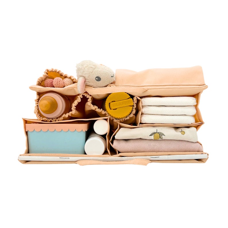 May include: A peach-colored diaper bag organizer with multiple compartments. The organizer is filled with baby essentials, including a bottle, a stuffed animal, a sippy cup, diapers, wipes, and a small container.