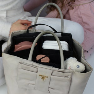 Pode incluir: Uma bolsa tote Prada bege com interior preto e um laço preto na frente. A bolsa está cheia de vários itens, incluindo uma tigela rosa, um pano branco e um animal de pelúcia branco.