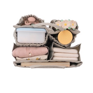 May include: A gray diaper bag with multiple compartments, filled with baby essentials. The bag contains a pacifier, a small container, a bottle of lotion, a stack of diapers, a baby outfit, and a book.