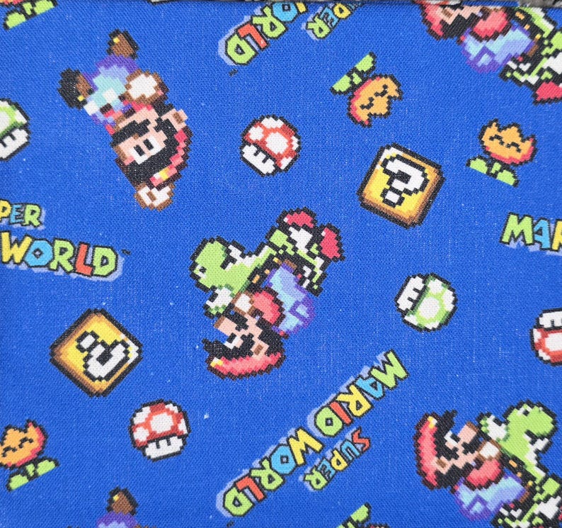May include: A blue fabric featuring a repeating pixelated pattern of Super Mario World characters including Mario riding Yoshi, red and white mushrooms, yellow question mark blocks, and orange flowers, with the text Super Mario World printed.