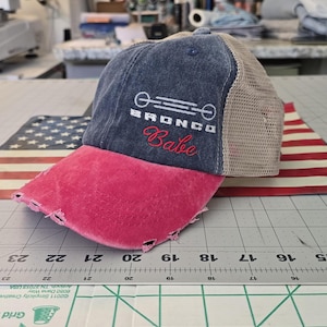 May include: A distressed baseball cap with a dark blue front panel, a bright pink frayed brim, and a tan mesh back. The front features a white graphic of a vehicle grille with the text "BRONCO" and the word "Babe" embroidered in red script.