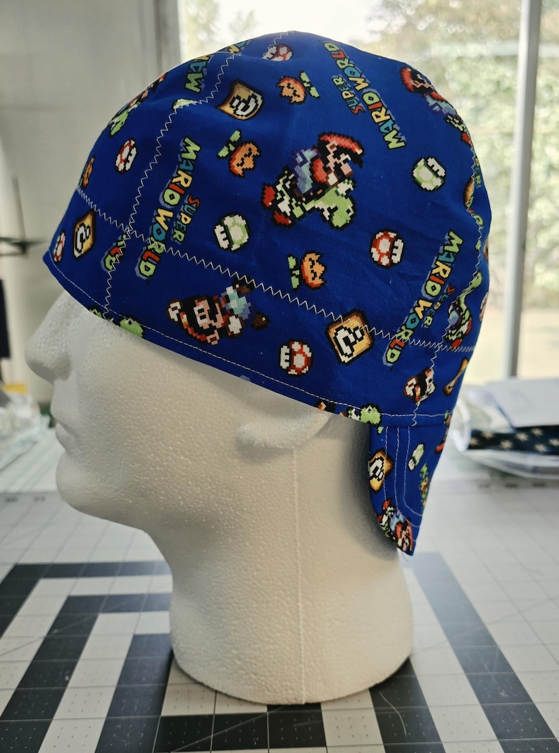 May include: A blue fabric cap featuring a repeating pixelated pattern of characters and items from Super Mario World, including Mario, Yoshi, and question blocks. The cap has visible white zigzag stitching along the seams and a short brim.
